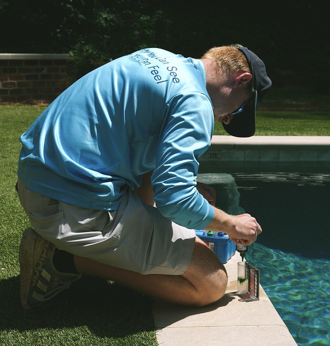 Man kneeling by a swimming pool edge, wearing a blue hoodie and cap, using a screwdriver on a small device placed on the pool deck.